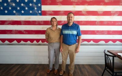 Amber Mordecai, new Executive Director of the Henryetta Chamber of Commerce, with Chamber President Kenny Wilson in front of an American flag wall