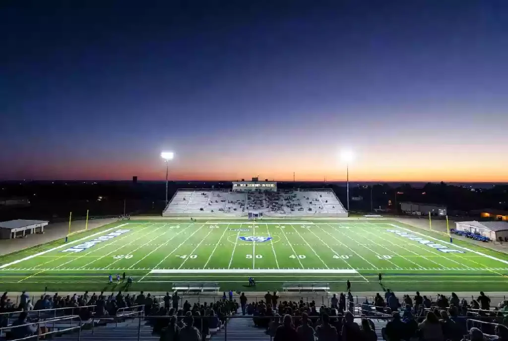 Wide view of a high school football stadium at dusk under stadium lights in Ada Oklahoma | Caption