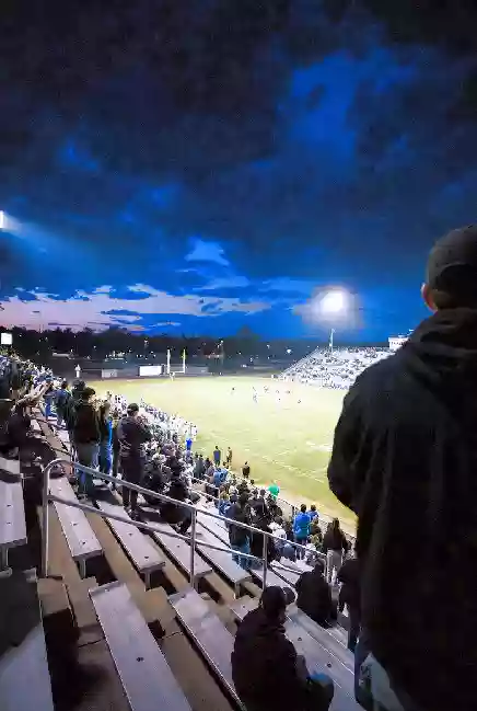 Vertical photo of fans watching a high school football game under the lights in Ada