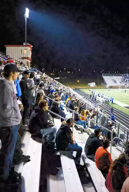 ertical photo of fans watching a high school football game under the lights in Mustang